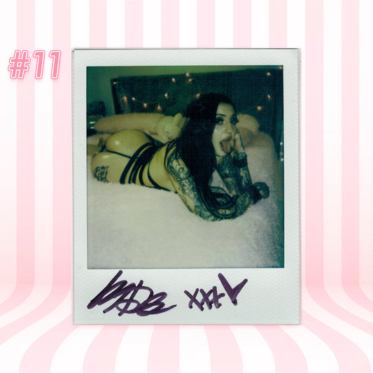 Polaroid-style photo of a person lying on a bed with decorative lights in the background, signed at the bottom.